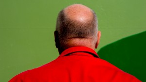 A man wearing a red jacket faces away from the camera in front of a green wall.
